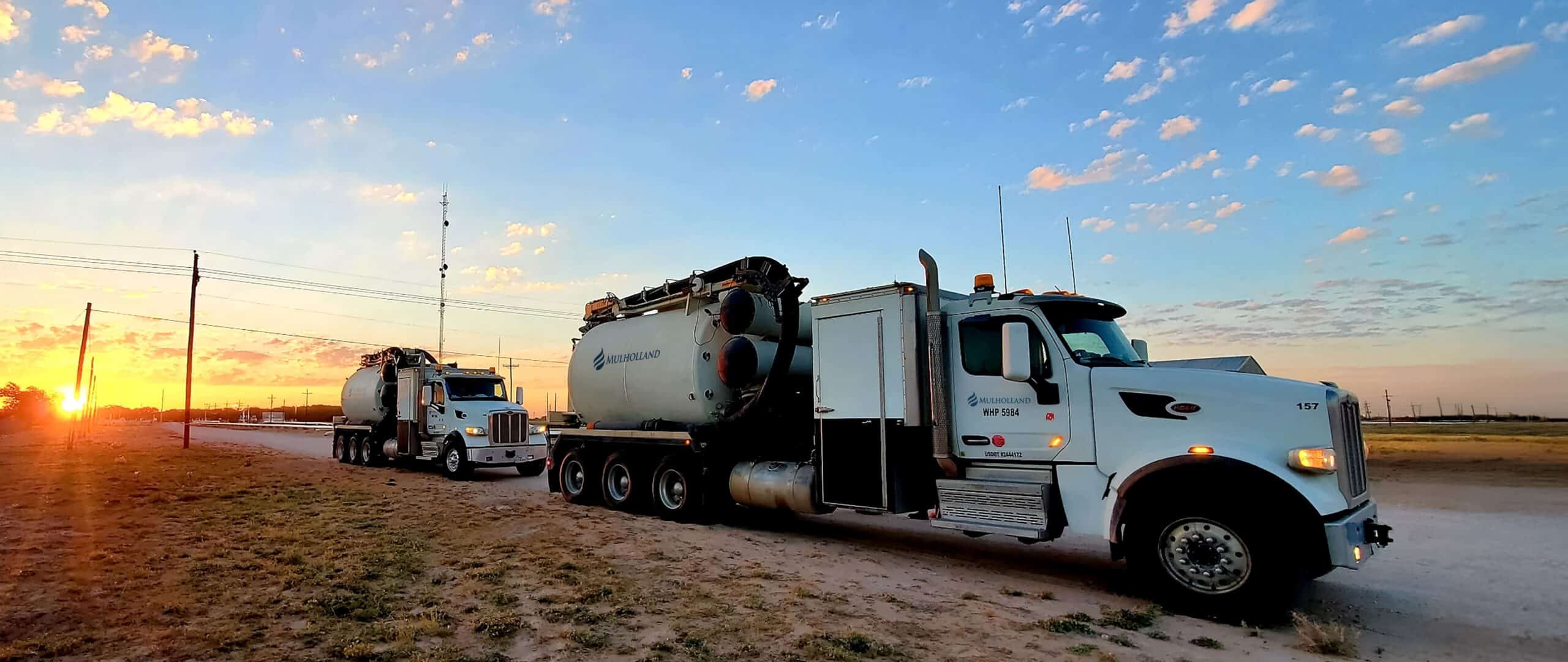 Two large trucks parked on a dirt road at sunset.