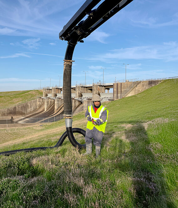 A man in a green vest standing next to a large pipe.